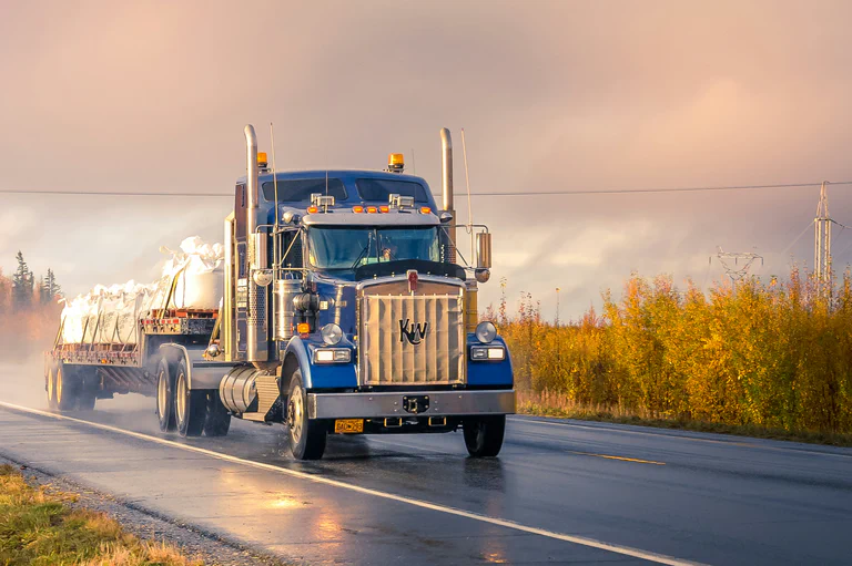 A photograph of a flatbed semi truck driving through a rural environment.